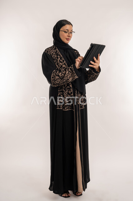تعابير الاندماج والتركيز، مفهوم إدارة المشاريع عن بعد، بورتريه لامرأة عربية خليجية اماراتية محجبة ترتدي العباءة السوداء تقوم بانجاز مهام العمل عبر جهاز التابلت، استخدام الاجهزة التقنية الحديثة, صورة الجسم بالطول الكامل, خلفية بيضاء