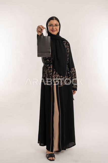 التسوق وطلب المنتجات عبر الإنترنت، مفهوم الاهتمام بالمظهر الخارجي, عملية تسليم الطلبات للزبائن، خدمات الشحن و التوصيل ذات الجودة العالية، بورتريه لامراة عربية خليجية اماراتية محجبة ترتدي العباءة السوداء تحمل بيدها كيس كرتون، خلفية بيضاء