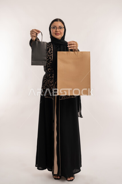 Concept of caring about appearance, process of delivering orders to customers, shopping and ordering products online, high quality shipping and delivery services, portrait of a veiled Arab Gulf Emirati woman wearing a black abaya holding bags in her hand, full body, white background