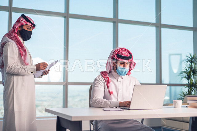 موظفان عربيان سعوديان خليجييان يعملان في  مقر الشركه , الالتزام بالاحترازات الوقائيه بارتداء الكمامه, متابعة ومناقشة الاعمال عبر جهاز اللابتوب , الوحده والعمل الجماعي, تحقيق اهداف الشركه, تطوير العمل