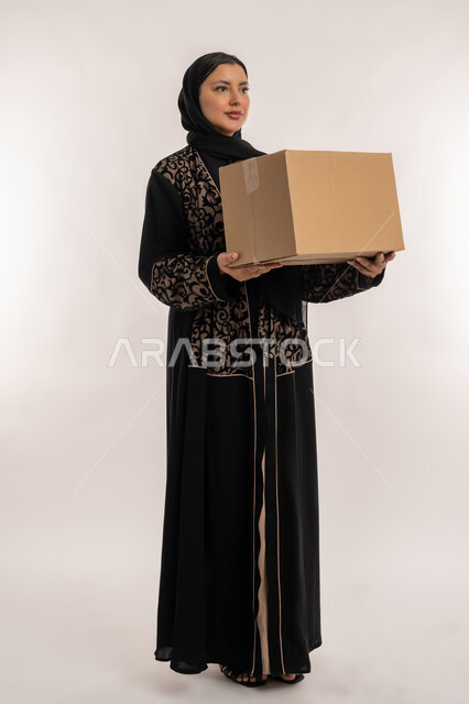 خدمات و الشحن التوصيل ذات الجودة العالية، عملية توصيل الطلبات للزبائن، التسوق و طلب المنتجات عبر الإنترنت، بورتريه لامراة عربية خليجية اماراتية محجبة ترتدي العباءة السوداء تحمل بيدها صندوق كرتوني، الجسم بالطول الكامل, خلفية بيضاء