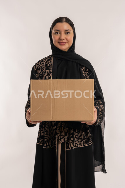 Looking at the camera with gestures of happiness and pleasure, high quality delivery services and shipping, customer delivery process, online shopping and ordering products, portrait of a veiled Emirati Gulf Arab woman wearing a black abaya holding a cardboard box, white background