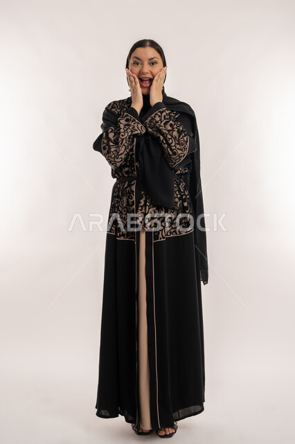 الاستغراب من شيء ما، تلقي اخبار مفرحة ومفاجئة، بورتريه لامراة عربية خليجية اماراتية محجبة ترتدي العباءة السوداء تضع يديها على خدها بإيماءات التعجب والصدمة والذهول, مفهوم الاهتمام بالمظهر الخارجي, الجسم بالطول الكامل, خلفية بيضاء