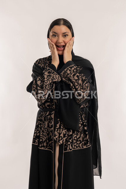 الاستغراب من شيء ما، تلقي اخبار مفرحة ومفاجئة، بورتريه لامراة عربية خليجية اماراتية محجبة ترتدي العباءة السوداء تضع يديها على خدها بإيماءات التعجب والصدمة والذهول, مفهوم الاهتمام بالمظهر الخارجي, خلفية بيضاء