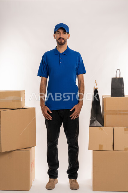 النظر الى الكاميرا بايماءات السعادة والفرح, إستلام المشتريات و تسليم البضائع للزبائن  بالوقت المحدد و بشكل سليم, بورتريه لمنوب توصيل عربي خليجي اماراتي يرتدي الزي الكاجوال و القبعة يحمل صندوق كرتوني, صورة الجسم بالطول الكامل, خلفيه بيضاء