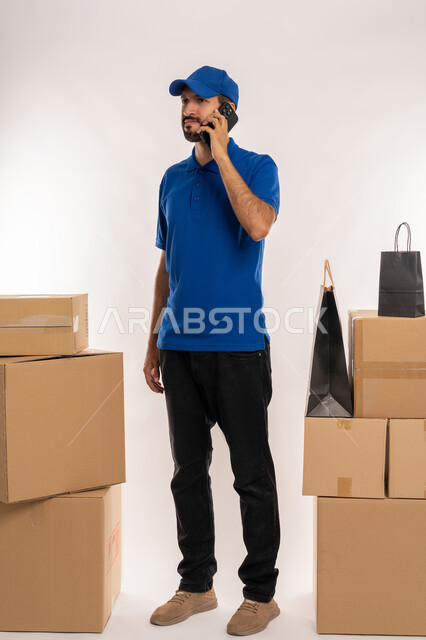 Making a phone call to confirm reservations and delivery times, integrating modern advanced technologies into work, shopping sites and centers and online shopping, portrait of an Arab Gulf Emirati delivery representative wearing casual clothes and a hat holding a cardboard box in his hand, full-length body, white background