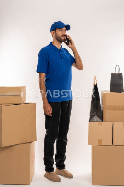 Making a phone call to confirm reservations and delivery times, integrating modern advanced technologies into work, shopping sites and centers and online shopping, portrait of an Arab Gulf Emirati delivery representative wearing casual clothes and a hat holding a cardboard box in his hand, full-length body, white background