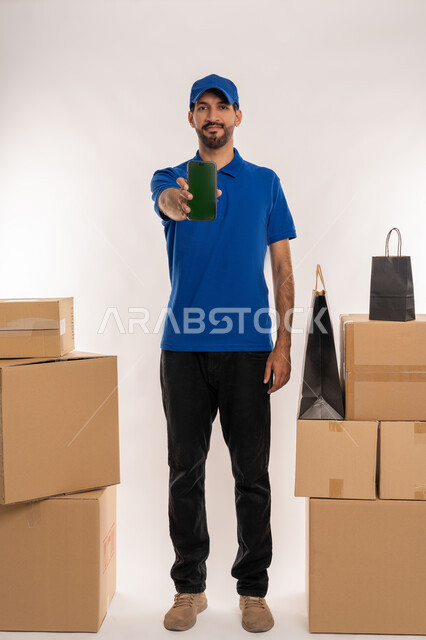 عرض شاشة سوداء فارغة عبر الهاتف المحمول, مواقع ومراكز التسوق والشراء الاونلاين, دمج التقنيات الحديثة المتطورة بالعمل، بورتريه لمندوب توصيل عربي خليجي اماراتي يرتدي الزي الكاجوال و القبعة يحمل بيده صنوق كرتوني، الجسم بالطول الكامل, خلفيه بيضاء