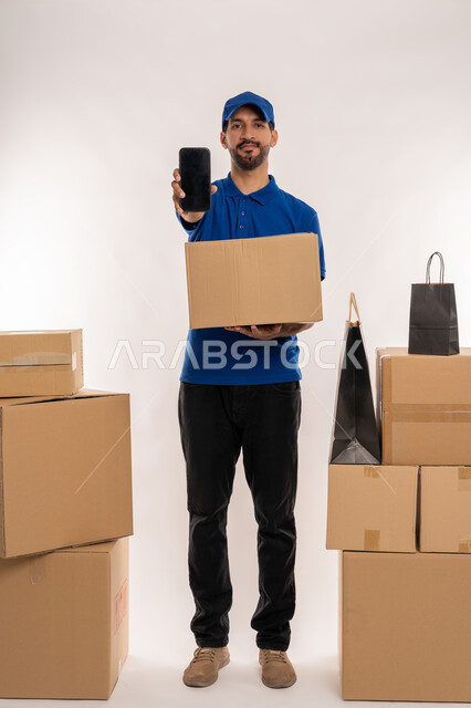 عرض شاشة سوداء فارغة عبر الهاتف المحمول, مواقع ومراكز التسوق والشراء الاونلاين, دمج التقنيات الحديثة المتطورة بالعمل، بورتريه لمندوب توصيل عربي خليجي اماراتي يرتدي الزي الكاجوال و القبعة يحمل بيده صنوق كرتوني، الجسم بالطول الكامل, خلفيه بيضاء