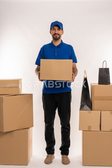 التسوق و طلب المنتجات عبر الإنترنت، إستلام المشتريات و تسليم البضائع للزبائن بالوقت المحدد و بشكل سليم، بورتريه لشاب عربي خليجي اماراتي يرتدي الكندورة و الطاقية الزرقاء يقف و يحمل بيده صندوق كرتوني، الجسم بالطول الكامل, خلفية بيضاء