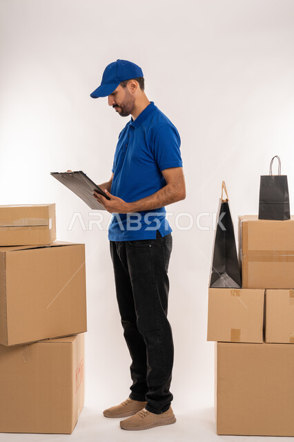 Writing daily tasks and goals, organizing and managing business, taking down important information and notes, office jobs and professions in the United Arab Emirates, portrait of a young Arab Gulf Emirati man wearing casual clothes and a blue hat holding a paper holder in his hand, full body, white background