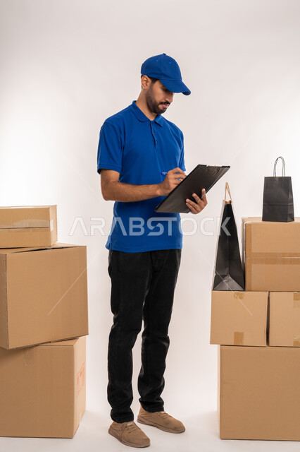 كتابة المهام والأهداف اليومية، تنظيم وإدارة الأعمال, تدوين المعلومات و الملاحظات المهمة، مهن ووظائف مكتبية في الامارات العربية المتحدة، بورتريه لشاب عربي خليجي اماراتي يرتدي الزي الكاجوال و القبعة الزرقاء يمسك بيده سنادة ورق, الجسم بالطول الكامل، خلفية بيضاء