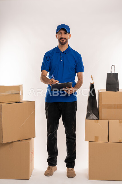 كتابة المهام والأهداف اليومية، تنظيم وإدارة الأعمال, تدوين المعلومات و الملاحظات المهمة، مهن ووظائف مكتبية في الامارات العربية المتحدة، بورتريه لشاب عربي خليجي اماراتي يرتدي الزي الكاجوال و القبعة الزرقاء يمسك بيده سنادة ورق, الجسم بالطول الكامل، خلفية بيضاء