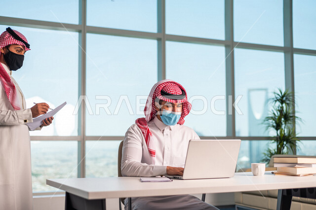 موظفان عربيان سعوديان خليجييان يعملان في  مقر الشركه , الالتزام بالاحترازات الوقائيه بارتداء الكمامه, متابعة ومناقشة الاعمال عبر جهاز اللابتوب , الوحده والعمل الجماعي, تحقيق اهداف الشركه, تطوير العمل