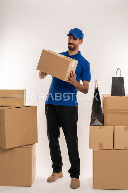 Shopping and ordering products online, receiving purchases and delivering goods to customers on time and in good condition, portrait of a young Arab Gulf Emirati man wearing a kandura and a blue cap standing and holding a cardboard box in his hand, full body, white background