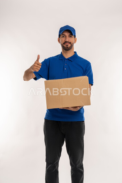 علامة الموافقة و الامتياز, خدمات الشحن والتوصيل ذات الجودة العالية, التسوق وطلب المنتجات عبر الإنترنت، بورتريه لشاب عربي خليجي اماراتي يرتدي الزي الكاجوال و القبعة الزرقاء يحمل صندوق كرتوني يرفع اصبع الابهام للاعلى بتعابير الرضا والقبول, خلفية بيضاء