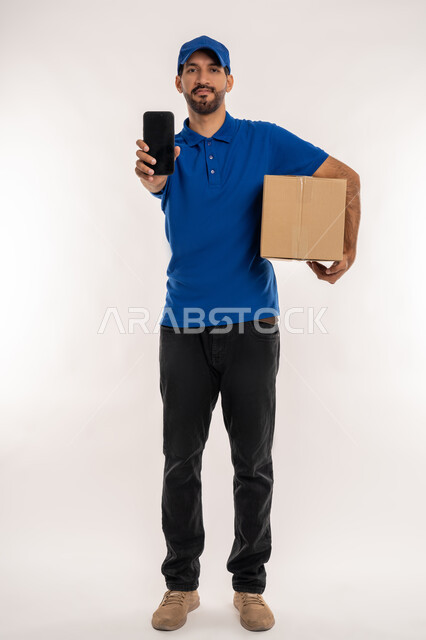 خدمات الشحن والتوصيل ذات الجودة العالية, التسوق وطلب المنتجات عبر الإنترنت، بورتريه لشاب عربي خليجي اماراتي يرتدي الزي الكاجوال و القبعة يحمل صندوق كرتوني, عرض شاشة سوداء فارغة عبر الجوال, استخدام التقنيات الحديثة المتطورة, الجسم بالطول الكامل, خلفية بيضاء