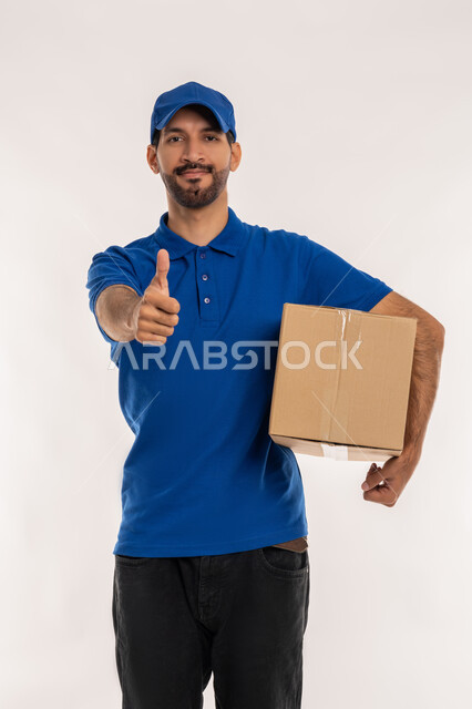 علامة الموافقة و الامتياز, خدمات الشحن والتوصيل ذات الجودة العالية, التسوق وطلب المنتجات عبر الإنترنت، بورتريه لشاب عربي خليجي اماراتي يرتدي الزي الكاجوال و القبعة الزرقاء يحمل صندوق كرتوني يرفع اصبع الابهام للاعلى بتعابير الرضا والقبول, خلفية بيضاء