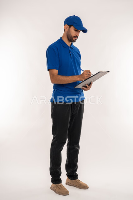 كتابة المهام والأهداف اليومية، تنظيم وإدارة الأعمال, تدوين المعلومات و الملاحظات المهمة، مهن ووظائف مكتبية في الامارات العربية المتحدة، بورتريه  لشاب عربي خليجي اماراتي يرتدي الزي الكاجوال و القبعة الزرقاء  يمسك بيده سنادة ورق ,الجسم بالطول الكامل، خلفية بيضاء