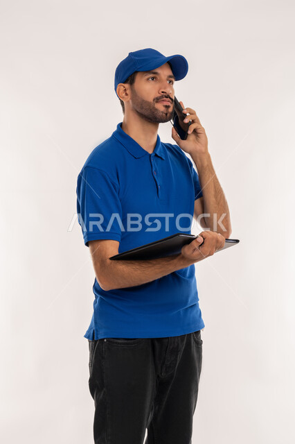 اجراء مكالمة صوتية عبر الهاتف المحمول، بورتريه  لشاب عربي خليجي اماراتي يرتدي الزي الكاجوال والقبعة الزرقاء يقوم بحمل جهاز التابليت, استخدام التقنيات الحديثة لإنجاز مهام العمل, إدارة شؤون الأعمال وتنظيمها, خلفية بيضاء