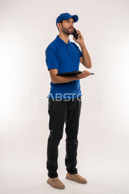 استخدام التقنيات الحديثة لإنجاز مهام العمل, اجراء مكالمة صوتية عبر الهاتف المحمول، بورتريه  لشاب عربي خليجي اماراتي يرتدي الزي الكاجوال والقبعة الزرقاء يقوم بحمل جهاز التابليت, إدارة شؤون الأعمال وتنظيمها, الجسم بالطول الكامل, خلفية بيضاء