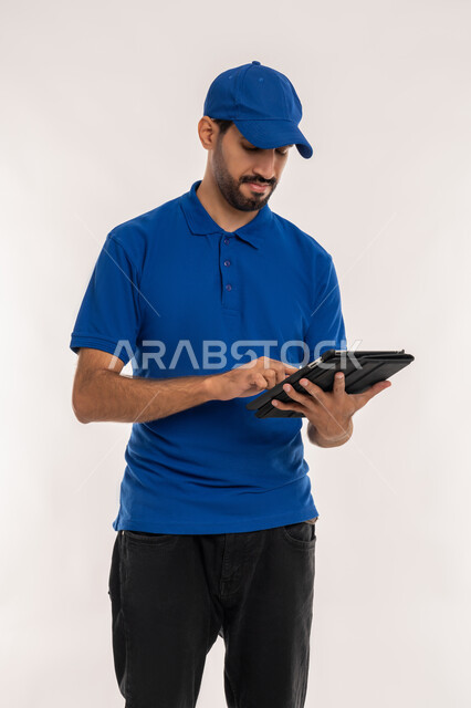 استخدام الأجهزة والتقنيات الحديثة, التواصل مع الاصدقاء عبر الانترنت, متابعه آخر أخبار السوشال ميديا, بورتريه  لشاب عربي خليجي اماراتي يرتدي الزي الكاجوال والقبعة الزرقاء يقوم بحمل جهاز التابليت, خلفية بيضاء