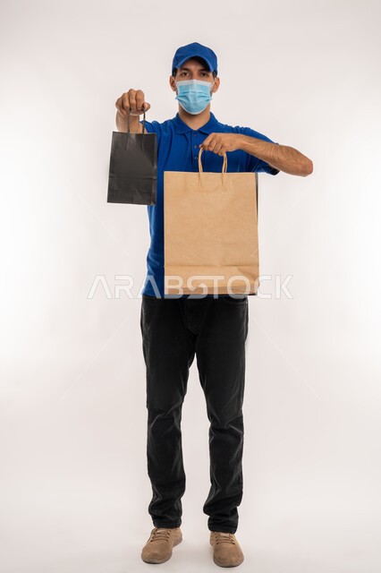 اتباع اجراءات الوقاية كوفيد 19, الاستفادة من العروض و التخفيضات, بورتريه لشاب عربي خليجي اماراتي يرتدي الزي الكاجوال والقبعة والكمامة ينظر الى الكاميرا و يحمل بيده اكياس التسوق , متعة في التسويق و اقتناء المستلزمات و الهدايا, الجسم بالطول الكامل, خلفيه بيضاء