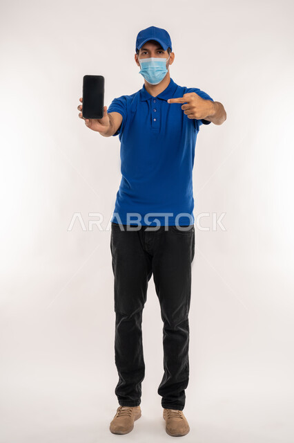اتباع الاجراءات الوقائية ضد فايروس كورونا, الالتزام بقوانين و قواعد وزارة الصحة, بورتريه لشاب عربي خليجي اماراتي يرتدي الزي الكاجوال والقبعة الزرقاء و الكمامة الوقائية يقوم بعرض شاشة سوداء فارغة على الهاتف المحمول ويشير بأصبع السبابة اليه, الجسم بالطول الكامل, خلفية بيضاء