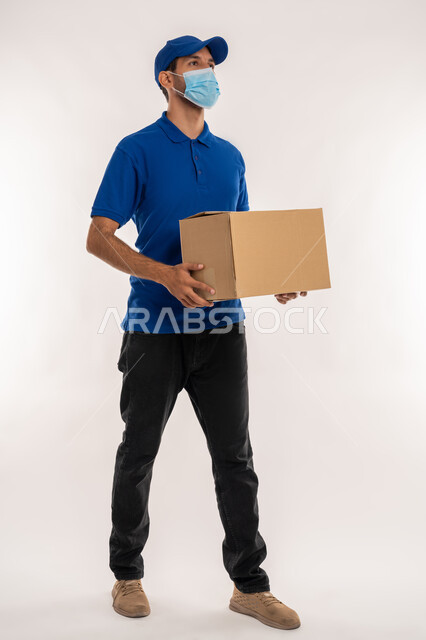 الالتزام بالاجراءات الاحترازية للوقاية من كوفيد 19, خدمات الشحن والتوصيل ذات الجودة العالية، التسوق وطلب المنتجات عبر الإنترنت، بورتريه لشاب عربي خليجي اماراتي يرتدي الزي الكاجوال و القبعة يحمل بيده صندوق ينظر الى شيء ما، الجسم بالطول الكامل, خلفية بيضاء