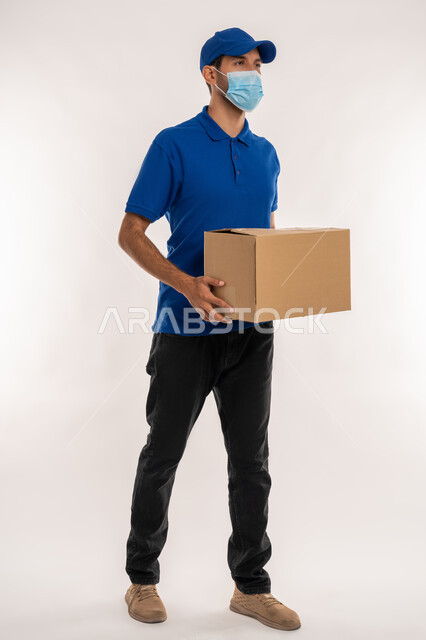 High quality shipping and delivery services, online shopping and ordering products, adherence to precautionary measures to prevent Covid-19, portrait of a young Arab Gulf Emirati man wearing casual clothes and a hat holding a box looking at something, full-length body, white background