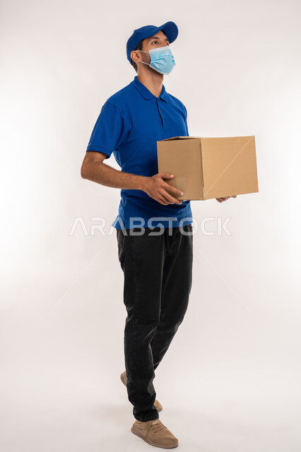 التسوق وطلب المنتجات عبر الإنترنت، خدمات الشحن والتوصيل ذات الجودة العالية، الالتزام بالاجراءات الاحترازية للوقاية من كوفيد 19, بورتريه لشاب عربي خليجي اماراتي يرتدي الزي الكاجوال و القبعة يحمل بيده صندوق ينظر الى شيء ما، الجسم بالطول الكامل, خلفية بيضاء