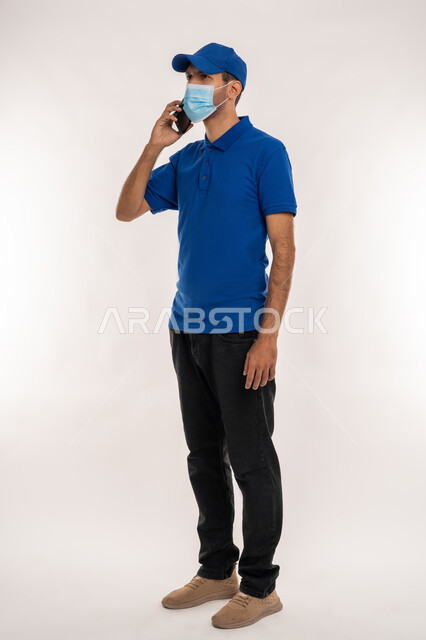 اتباع الاجراءات الوقائية ضد فايروس كورونا, الالتزام بقوانين و قواعد وزارة الصحة لتجنب الاصابه بكوفيد 19, بورتريه لشاب عربي خليجي اماراتي يرتدي الزي الكاجوال و القبعة والكمامة الوقائية يقوم باجراء مكالمة هاتفية عبر الهاتف المحمول, الجسم بالطول الكامل, خلفية بيضاء