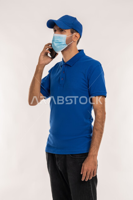 الالتزام بقوانين و قواعد وزارة الصحة لتجنب الاصابه بكوفيد 19, اتباع الاجراءات الوقائية ضد فايروس كورونا, بورتريه لشاب عربي خليجي اماراتي يرتدي الزي الكاجوال والقبعة الزرقاء و الكمامة الوقائية يقوم باجراء مكالمة هاتفية عبر الهاتف المحمول, خلفية بيضاء