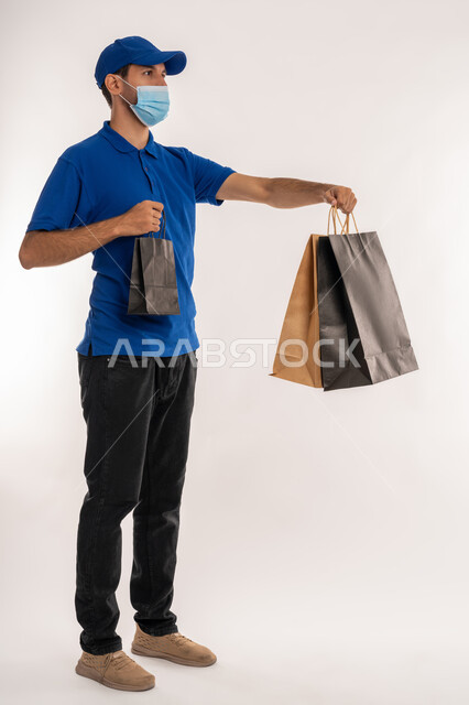 Benefit from offers and discounts, follow Covid-19 prevention measures, portrait of a young Arab Gulf Emirati man wearing casual clothes, hat and mask, looking at the camera and holding shopping bags, fun in marketing and purchasing supplies and gifts, full body, white background