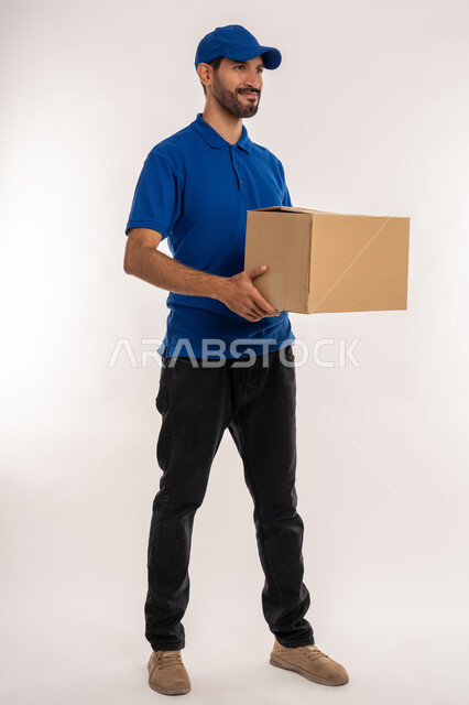 عملية تسليم الطلبات للزبائن، التسوق وطلب المنتجات عبر الإنترنت، خدمات الشحن و التوصيل ذات الجودة العالية، بورتريه لشاب عربي خليجي اماراتي يرتدي الالزي الكاجوال و قبعة زرقاء يحمل بيده أكياس، الجسم بالطول الكامل, خلفية بيضاء