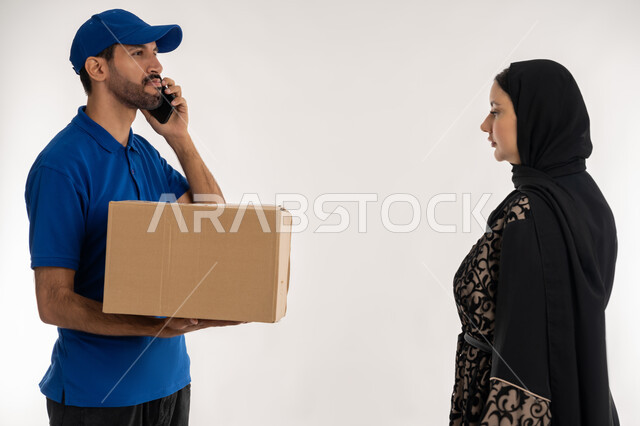Online shopping and shopping malls, integrating advanced modern technologies into work, portrait of a young Arab Gulf Emirati man wearing casual clothes and a blue hat delivering orders standing next to an Arab Gulf Emirati woman, making a phone call to confirm reservations and delivery times, white background