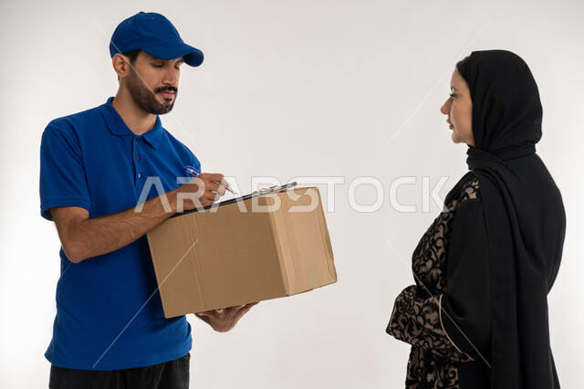 دمج العمل مع التقنية والتكنولوجيا، شبكات التوصيل الاونلاين في الخليج، التسوق وطلب المنتجات عبر الإنترنت, بورتريه لشاب عربي خليجي اماراتي يرتدي الزي الكاجوال و القبعة الزرقاء يقوم بتوثيق استلام الطرود يقف بجانب امرأة عربية خليجية امارتيه ، خلفيه بيضاء