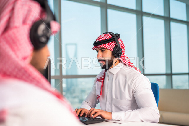موظفان عربيان سعوديان خليجيان يعملان في خدمة العملاء في شركه يرتديان سماعة الاتصال, استعلامات واستفسارات, الاجابه على اسئلة العملاء, اجراء اتصال فيديو من خلال الحاسوب , عقد اجتماع عمل عبر الانترنت, العمل عن بعد , متابعة وتطوير العمل .