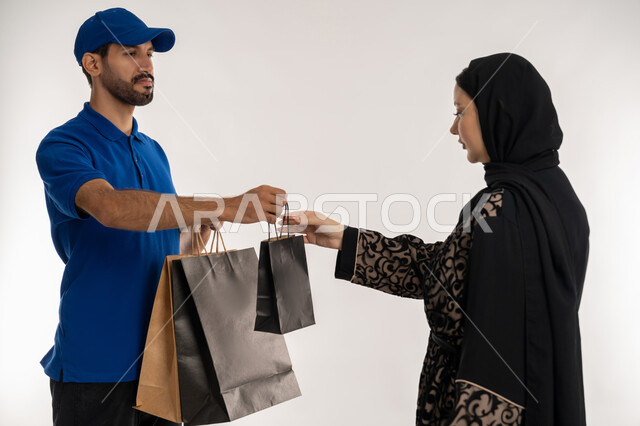 التسوق وطلب المنتجات عبر الإنترنت, شبكات التوصيل الاونلاين في الخليج، دمج العمل مع التقنية والتكنولوجيا، بورتريه لشاب عربي خليجي اماراتي يرتدي الزي الكاجوال و القبعة الزرقاء يقف بجانب امرأة عربية خليجية امارتيه يقوم بتوصيل الطلبات، خلفيه بيضاء