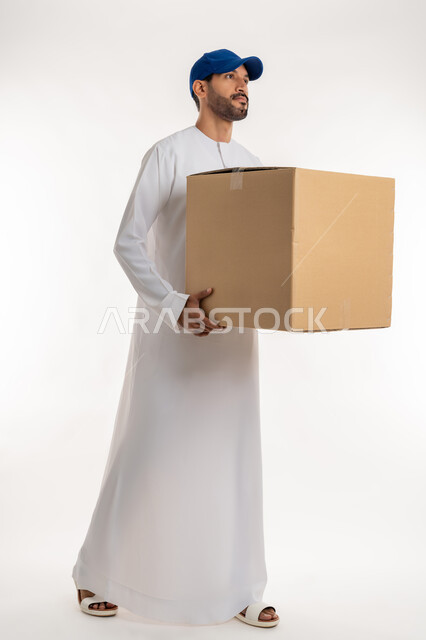 التسوق وطلب المنتجات عبر الإنترنت، عملية توصيل الطلبات للزبائن، خدمات الشحن و التوصيل ذات الجودة العالية، بورتريه لشاب عربي خليجي اماراتي يرتدي الكندورة و الطاقية الزرقاء يحمل بيده صندوق كرتوني، الجسم بالطول الكامل, خلفية بيضاء