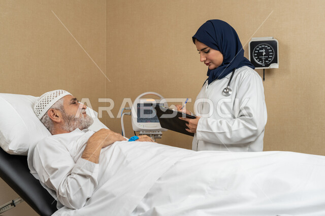 تقديم الخدمات والاستشارات الطبية للمراجعين، مفهوم الطب والرعاية الصحية، طبيبة عربية خليجية اماراتية ترتدي الحجاب والمعطف الابيض تقوم بتدوين معلومات مريض مسن عربي اماراتي مستلقي على السرير، مهن ووظائف نسائية، دراسة و مراجعة حالات المرضى