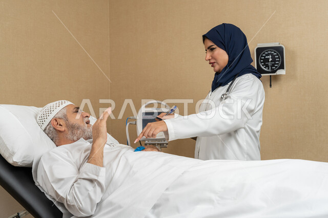 تقديم الخدمات والاستشارات الطبية للمراجعين، مفهوم الطب والرعاية الصحية، طبيبة عربية خليجية اماراتية ترتدي الحجاب والمعطف الابيض تقوم بتدوين معلومات مريض مسن عربي اماراتي مستلقي على السرير، مهن ووظائف نسائية، دراسة و مراجعة حالات المرضى