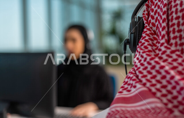 صوره مقربه  لخلفية رأس موظف  عربي سعودي خليجي في مقر العمل يرتدي  سماعة الاتصال والشماغ السعودي  . زملاء عمل في مركز الاستعلامات, اجراء اتصال فيديو من خلال الحاسوب , عقد اجتماع عمل عبر الانترنت , العمل عن بعد , متابعة وتطوير العمل.