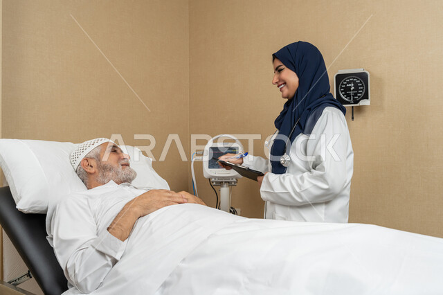 تقديم الخدمات والاستشارات الطبية للمراجعين، مفهوم الطب والرعاية الصحية، طبيبة عربية خليجية اماراتية ترتدي الحجاب والمعطف الابيض تقوم بتبادل الاحاديث والمعلومات مع مريض مسن عربي اماراتي مستلقي على السرير، مهن ووظائف نسائية، دراسة و مراجعة حالات المرضى