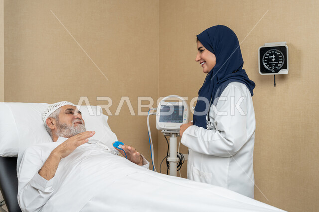 تقديم الخدمات والاستشارات الطبية للمراجعين، مفهوم الطب والرعاية الصحية، طبيبة عربية خليجية اماراتية ترتدي الحجاب والمعطف الابيض تقوم بتبادل الاحاديث والمعلومات مع مريض مسن عربي اماراتي مستلقي على السرير، مهن ووظائف نسائية، دراسة و مراجعة حالات المرضى