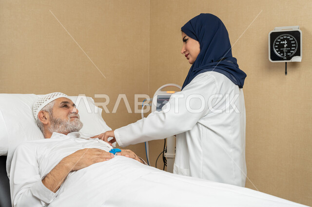 تقديم الخدمات والاستشارات الطبية للمراجعين، مفهوم الطب والرعاية الصحية، طبيبة عربية خليجية اماراتية ترتدي الحجاب والمعطف الابيض تقوم بتبادل الاحاديث والمعلومات مع مريض مسن عربي اماراتي مستلقي على السرير، مهن ووظائف نسائية، دراسة و مراجعة حالات المرضى
