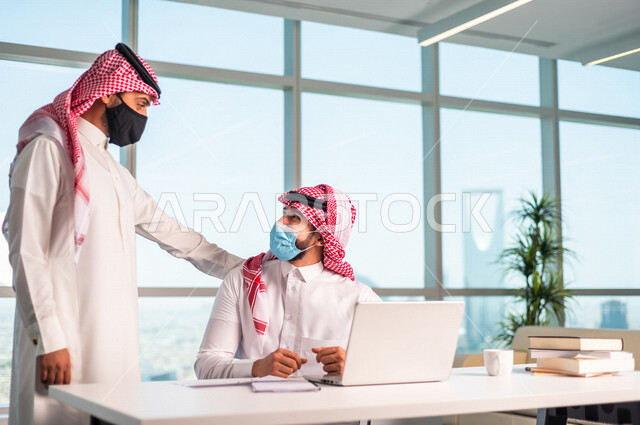 موظفان عربيان سعوديان خليجييان يعملان في  مقر الشركه , الالتزام بالاحترازات الوقائيه بارتداء الكمامه, متابعة ومناقشة الاعمال عبر جهاز اللابتوب , الوحده والعمل الجماعي, تحقيق اهداف الشركه, تطوير العمل