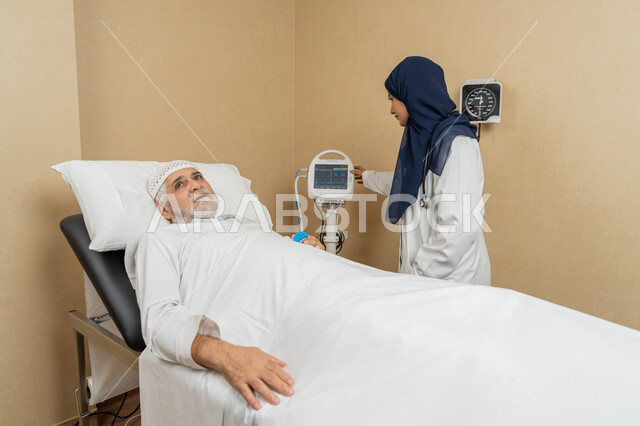 تقديم الخدمات والاستشارات الطبية للمراجعين، مفهوم الطب والرعاية الصحية، طبيبة عربية خليجية اماراتية ترتدي الحجاب والمعطف الابيض تقوم بتبادل الاحاديث والمعلومات مع مريض مسن عربي اماراتي مستلقي على السرير، مهن ووظائف نسائية، دراسة و مراجعة حالات المرضى
