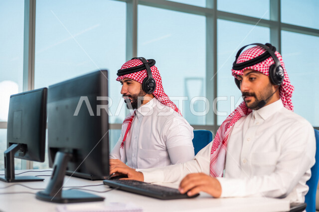 موظفان عربيان سعوديان خليجيان يعملان في خدمة العملاء في شركه يرتديان سماعة الاتصال, استعلامات واستفسارات, الاجابه على اسئلة العملاء, اجراء اتصال فيديو من خلال الحاسوب , عقد اجتماع عمل عبر الانترنت, العمل عن بعد , متابعة وتطوير العمل .