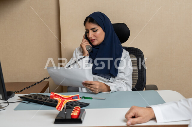 تقديم الخدمات الطبية للمرضى بكل كفاءة، مهن ووظائف نسائية، صورة مقربة لطبية اماراتية عربية خليجية تجلس خلف مكتبها تجري مكالمة هاتفية للاستعلام عن أمر ما، مفهوم الطب والرعاية الصحية